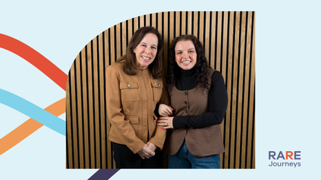 Two people standing close together with arms linked, smiling, in front of a wood slat wall.